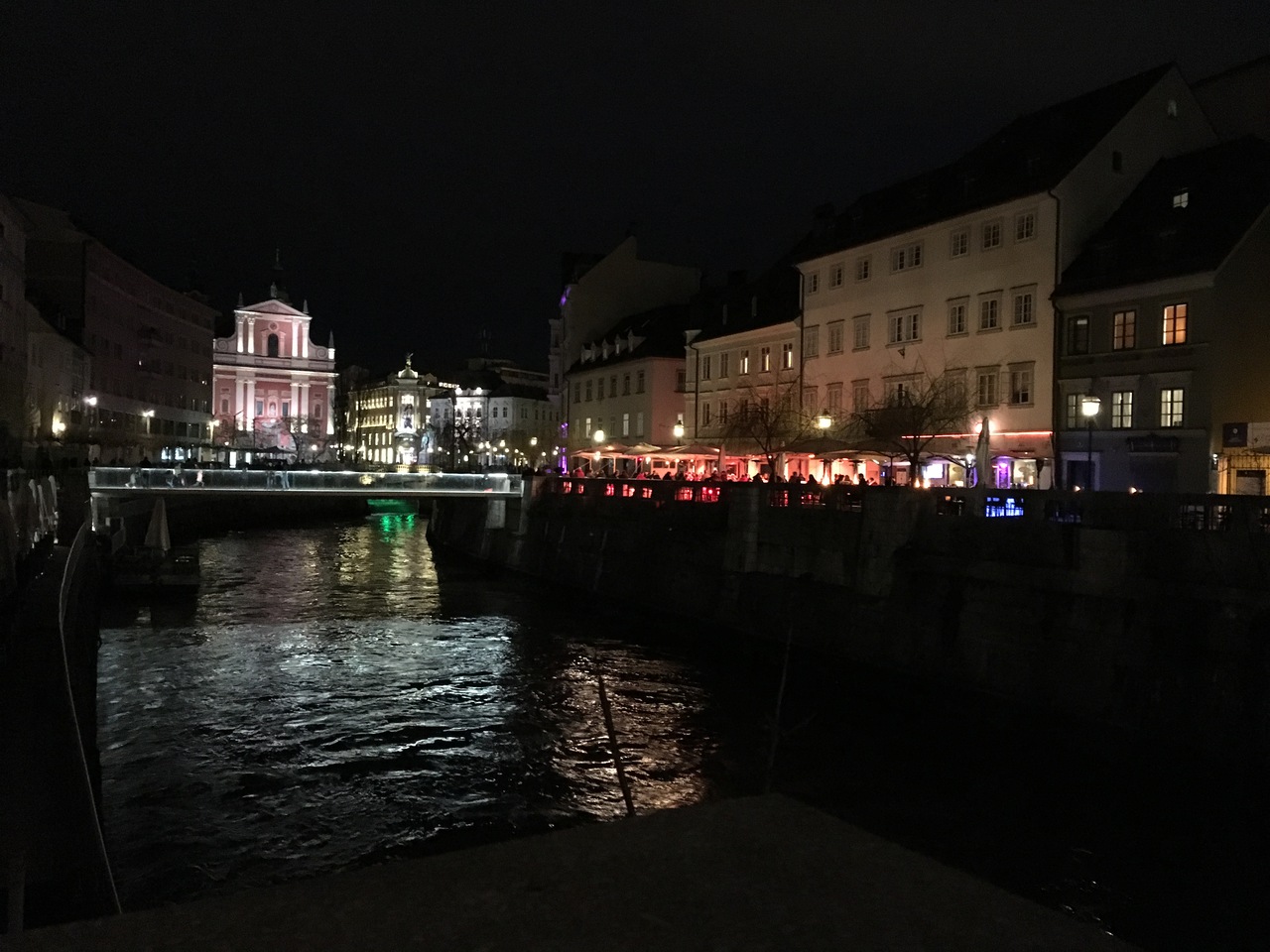 Shot across the river, people sitting around heating lamps in front of a brightly lit bar, shot taken during the dark of the night