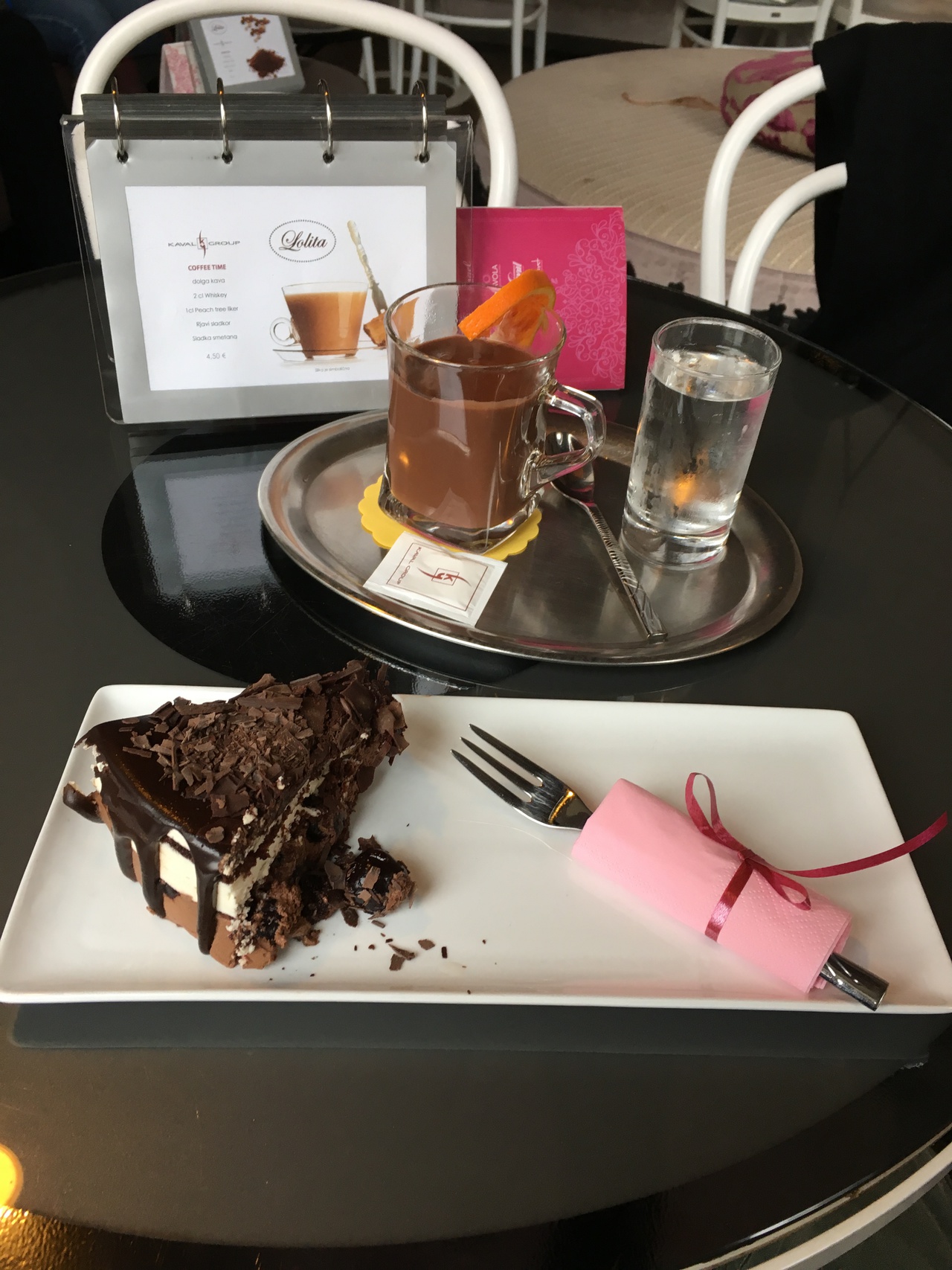 A rectangular dish with a small piece of Black Forest cake. Behind the dish a cup with liquid brown chocolate